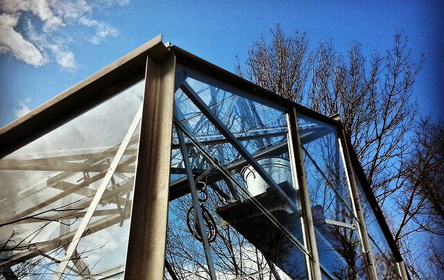 Estructura de vidrio y metal de un invernadero vista desde abajo, con reflejos en los cristales y árboles sin hojas al fondo bajo un cielo azul.