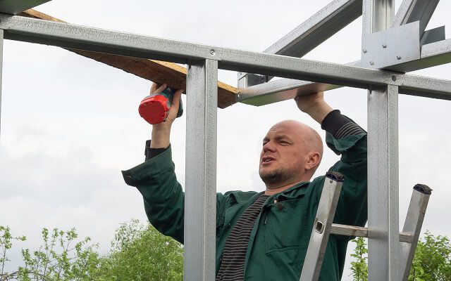 Hombre utilizando un taladro para fijar una estructura metálica con una pieza de madera, mientras trabaja en un marco al aire libre.